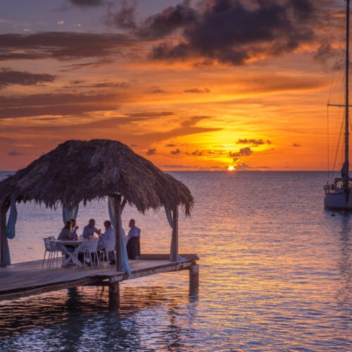 Sunset over a beach restaurant in Bonaire, tables and chairs, and the ocean in the background under a colorful orange and pink sky.